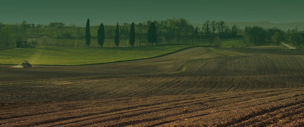 Paysage agricole vallonné: champs labourés bruns, prairies vertes, tracteur cultivant, bâtiments ruraux et arbres au loin.